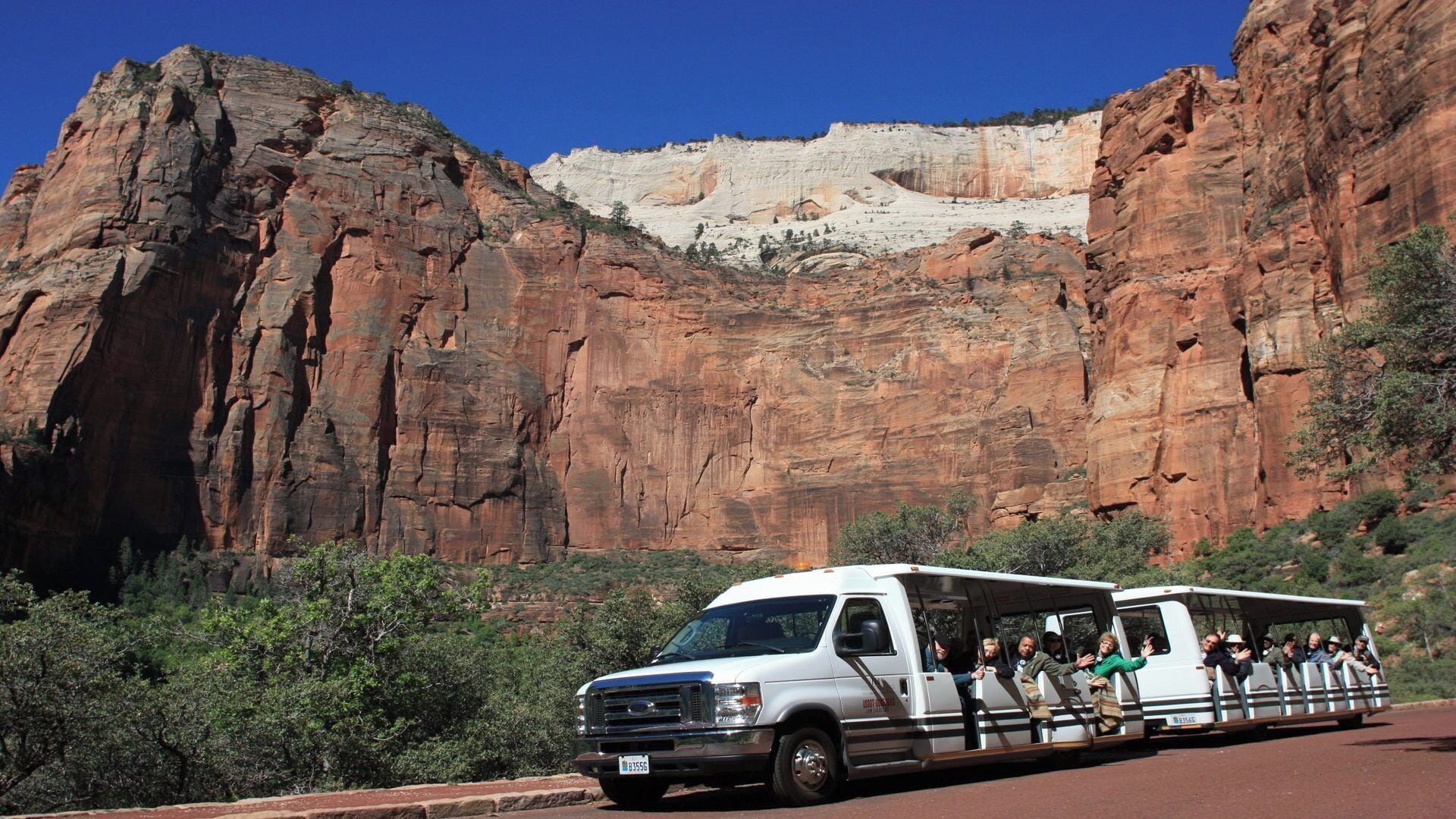 A white tour bus with passengers, parked in front of a large red rock formation.