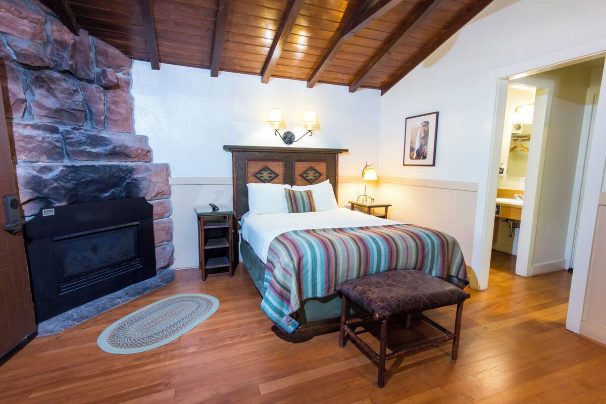 Un dormitorio con chimenea de piedra, una cama con cubrecama a rayas, un banco de madera y una mesita de noche con lámpara.