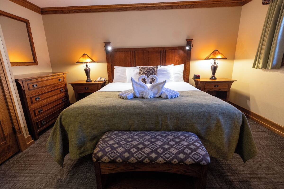 Una cama bien tendida con una decoración de toallas, una mesita de noche de madera con lámpara y un banco con cojín estampado.