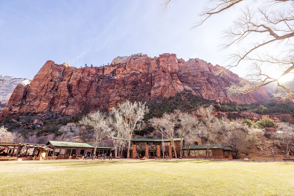 A building with a green roof, surrounded by a grassy area and trees, with a large red rock formation in the background. - Zion Lodge - Inside the Park (Springdale)