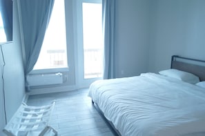 A bedroom with a bed, a window with curtains, a radiator, and a white chair.