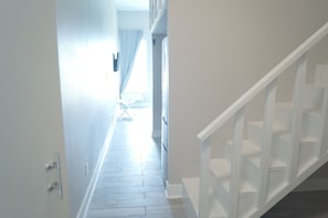 A narrow hallway with a staircase on the right, a door at the end, and a window with curtains.