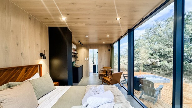 Una camera da letto moderna con un ampio letto, una testiera in legno, un angolo cottura e vista sull'esterno attraverso ampie finestre.