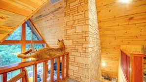 Interior de una cabaña de madera con pared de piedra, una escalera de madera y un gato en la barandilla.