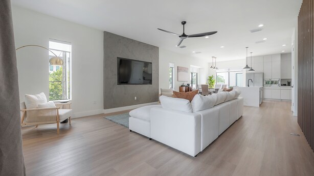 Un salon moderne avec un grand canapé d’angle blanc, un téléviseur à écran plat fixé au mur et un ventilateur de plafond.