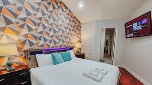 A modern hotel room with a geometric-patterned wall, a large bed with white linens, two bedside tables with lamps, and a flat-screen TV mounted on the wall. - Walnut Premium / King Bed / 65" TV / Ping-Pong (Philadelphia)