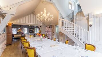 Une salle à manger avec une longue table dressée pour un repas, un lustre et un escalier menant à un niveau supérieur.