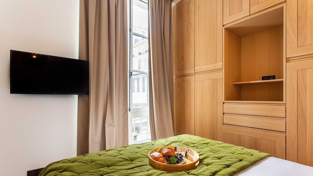 A hotel room with a bed, a flat-screen TV mounted on the wall, a wooden wardrobe, and a window with curtains. - Comfortable Apartment - 1br/2p - Le Marais (Paris)