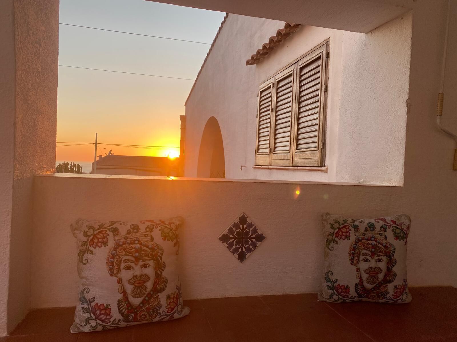 A balcony with two decorative pillows, a window with shutters, and a sunset view.