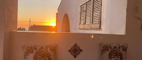 A balcony with two decorative pillows, a window with shutters, and a sunset view.