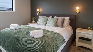 A neatly made bed with a green blanket and two rolled towels, a bedside table with a lamp, and a small plant on the nightstand.