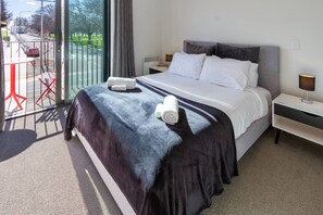 A hotel room with a large bed, a nightstand, a lamp, and a view of a parking area and greenery outside. - Stadium Edge Retreat  Modern 2 Bed with Carpark (Christchurch)