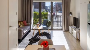 Ein modernes Wohnzimmer mit einer Couch, einem kleinen Tisch mit einer Vase, einem Sessel, einem Fernseher und einer Glastür, die zu einem Außenbereich führt.