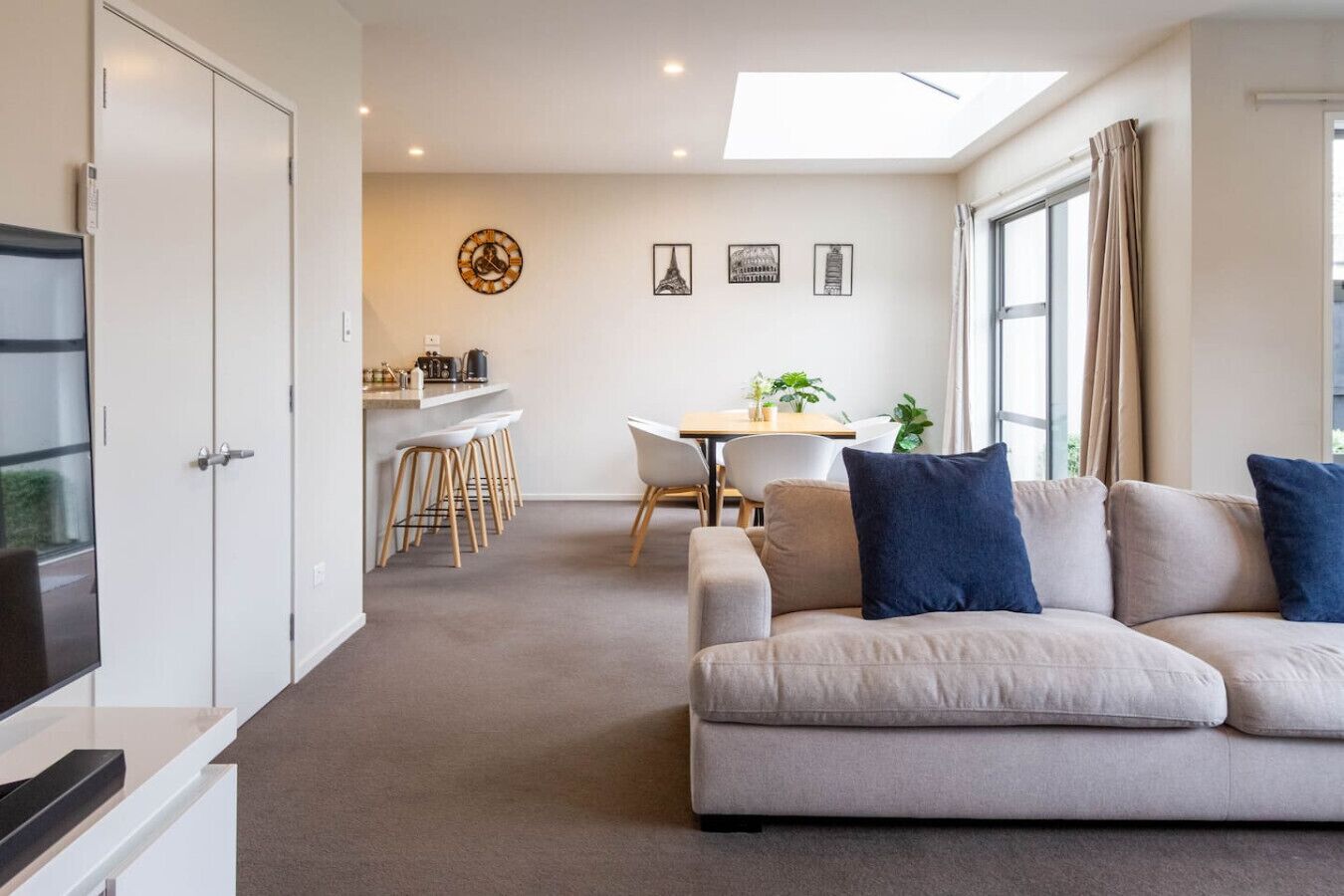 Ein modernes Wohnzimmer mit einer Couch, einem Essbereich mit Tisch und Stühlen sowie einer Küchenzeile mit einer Arbeitsplatte und Hockern.