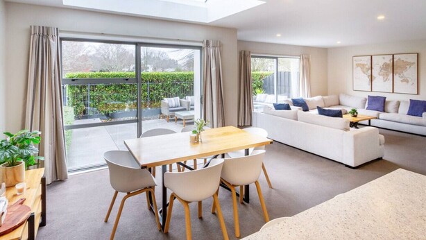 A modern living room with a large sofa, a dining table with chairs, and a balcony with a view of greenery.