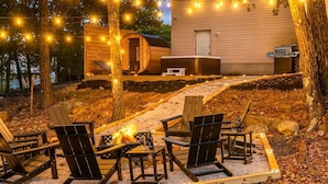 A cozy outdoor seating area with Adirondack chairs and a fire pit, surrounded by a wooden deck and string lights.