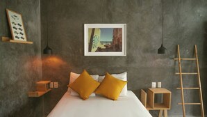 A bedroom with a bed, two mustard-colored pillows, a framed picture of a coastal scene, a small wooden shelf, and a ladder against the wall.