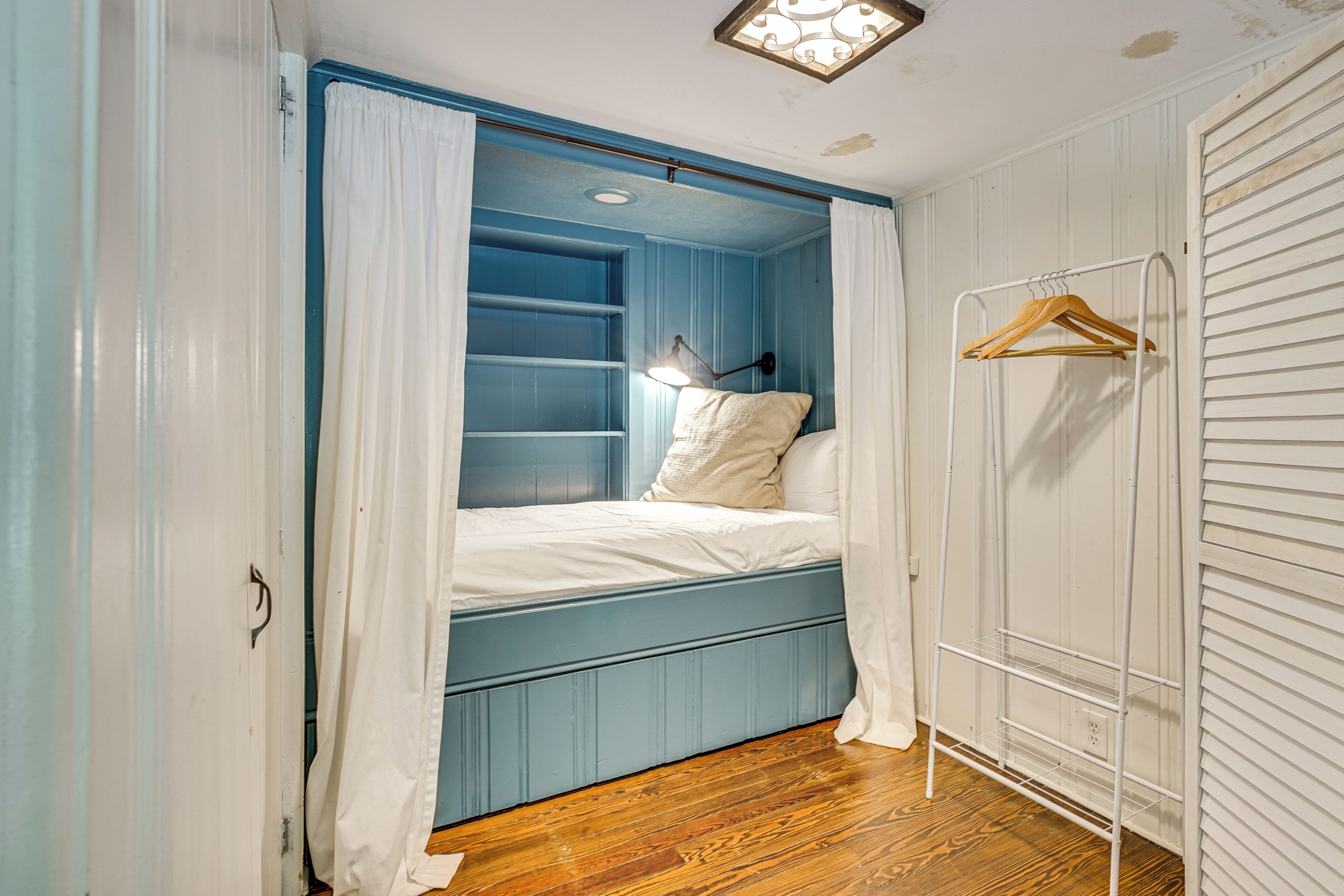 A bedroom with a blue bed, white curtains, a bedside lamp, and a wooden hanger rack.