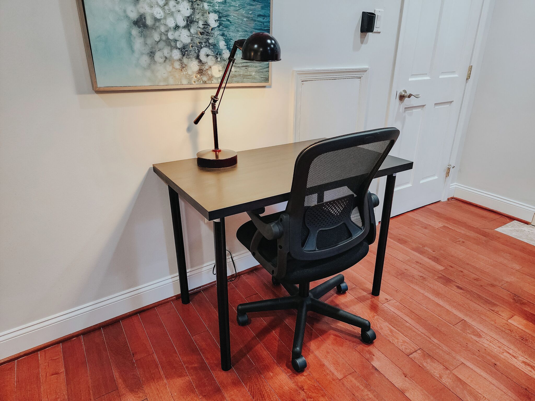 A small office space with a wooden desk, a black office chair, a black desk lamp, and a painting on the wall.