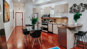 Una cocina moderna con electrodomésticos de acero inoxidable, un comedor con mesa de vidrio y sillas negras, y una sala de estar con piso de madera y una planta grande.
