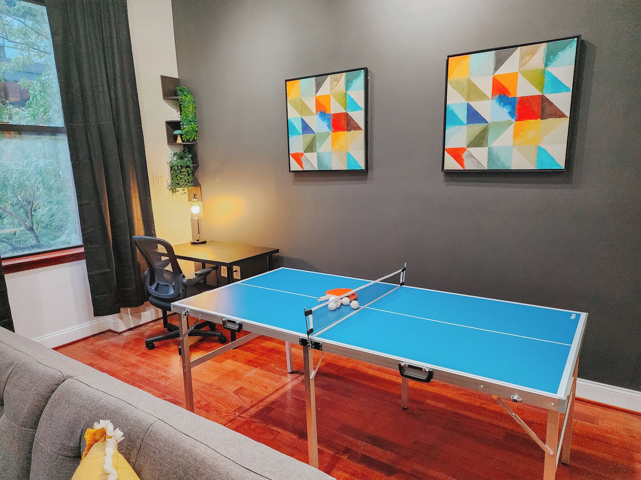 A room with a ping pong table, a gray sofa, and two abstract paintings on the wall.