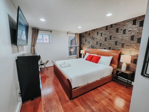 Habitación de hotel con piso de madera, cama con almohadas blancas y rojas, televisor montado en la pared y una pared con diseño.