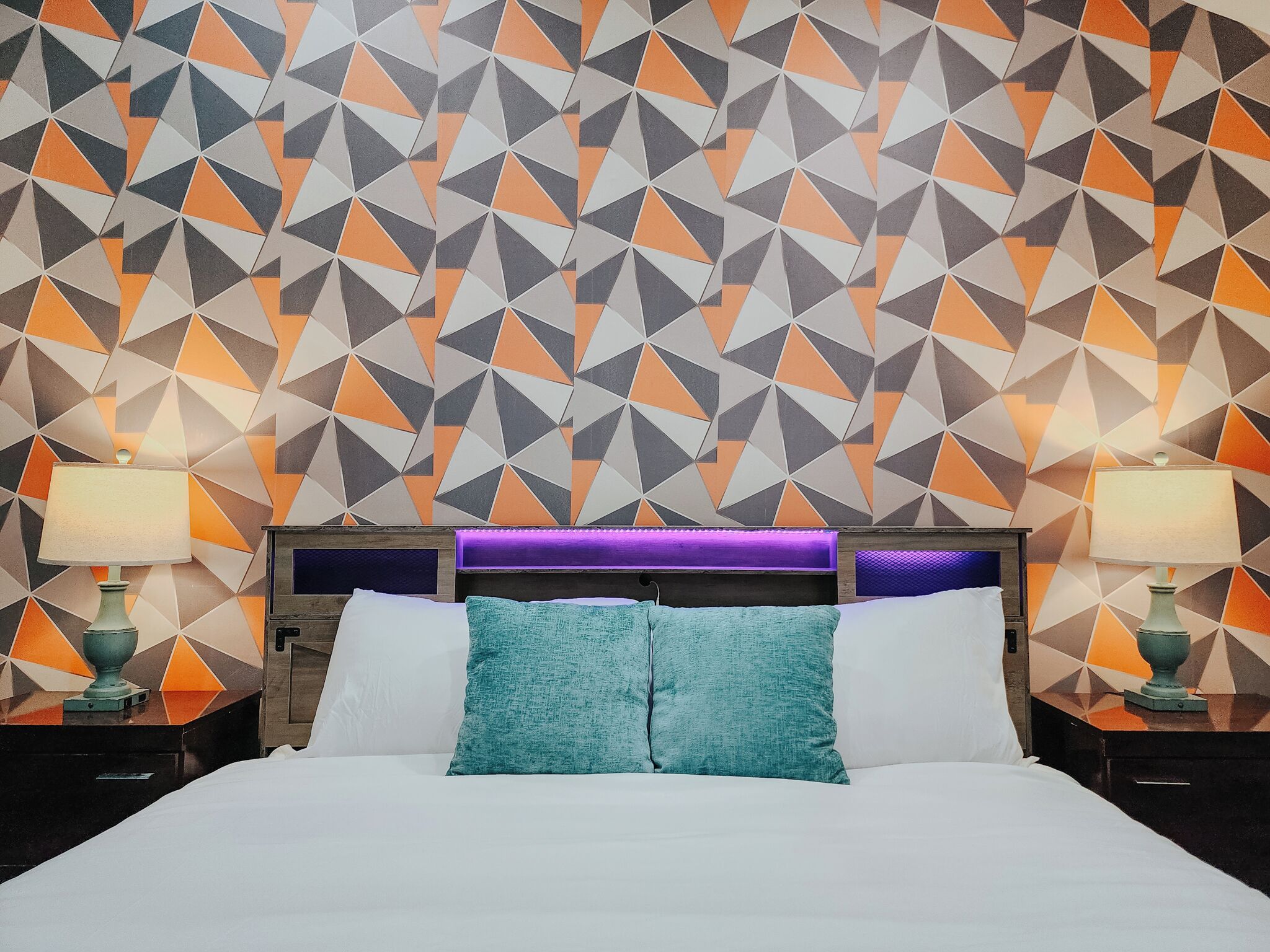 Une chambre d’hôtel avec un mur orné d’un motif géométrique, un lit avec du linge de lit blanc, des coussins turquoise et deux tables de chevet avec des lampes.