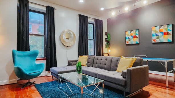 Una sala de estar moderna con un sofá gris, un sillón azul, una mesa de centro de vidrio y una mesa de ping pong.