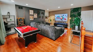Una sala de estar moderna con una mesa de billar, un sofá seccional gris, un televisor de pantalla plana y una pared decorativa con plantas.
