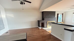 A spacious room with a wooden floor, a ceiling fan, a stone fireplace, and a kitchen area with white cabinets. - Spacious 3-bedroom Lakeview Home Centrally Located in Quiet Neighborhood (Moses Lake)