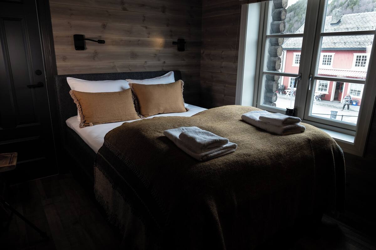 Une chambre à coucher avec une tête de lit en bois, une grande fenêtre donnant sur un immeuble rouge, et un lit bien fait avec deux oreillers et des serviettes pliées.