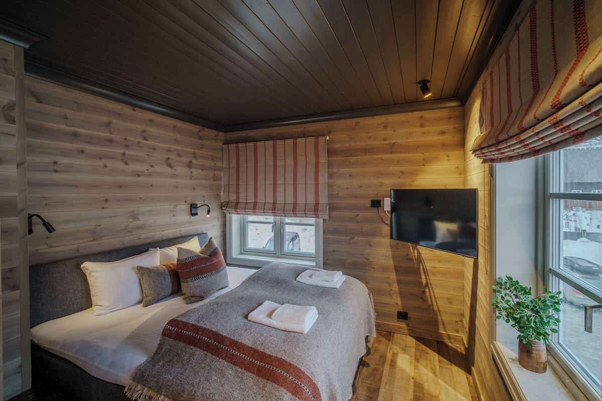 Une chambre confortable au décor intérieur en bois, avec un grand lit recouvert d’oreillers, une fenêtre avec des stores et une petite plante posée sur le rebord de la fenêtre.