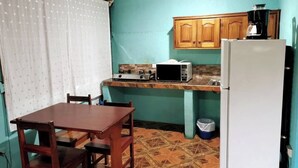 A compact kitchen with a microwave, stove, and refrigerator. There is a wooden table and chairs, and a window with curtains.
