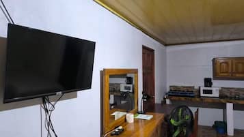 A room with a flat-screen TV mounted on the wall, a wooden desk with a chair, a microwave, and a small cabinet.