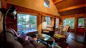 A cozy cabin interior with a sofa, armchair, and a glass-top coffee table. Large windows offer views of the outdoors.