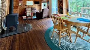 A spacious wooden cabin with a high ceiling, a fireplace, a dining area with a round table and chairs, and a large window offering a view of the outdoors.