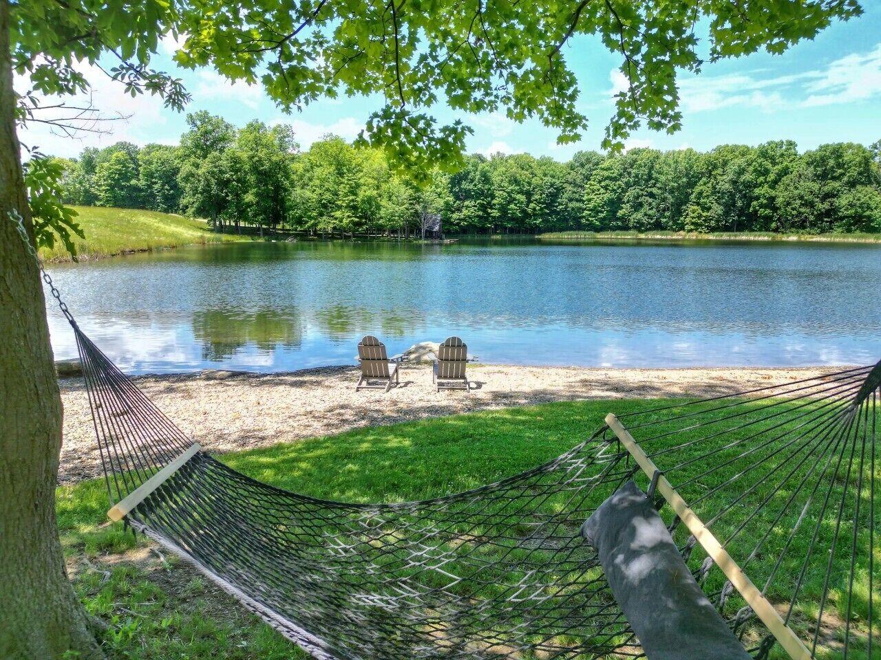 En hängmatta och två Adirondack-stolar vid en sjö, med en skog i bakgrunden.
