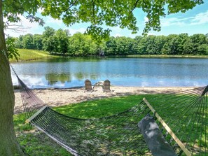 A hammock and two Adirondack chairs by a lake with a forest backdrop.