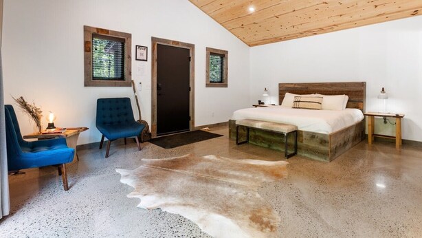 Un dormitorio con techo de madera, una cama con cabecera de madera, una silla azul, un banco y una alfombra de cuero de vaca.