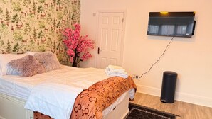 Une chambre à coucher avec un lit, un téléviseur fixé au mur, une porte et une poubelle.