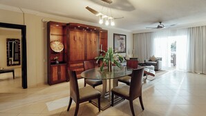 A spacious dining area with a round glass table and four chairs, a wooden cabinet with decorative items, and a ceiling fan.