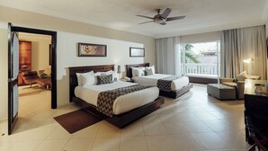 A hotel room with two beds, a ceiling fan, a large window with curtains, a seating area with a sofa and armchair, and a desk with a lamp.