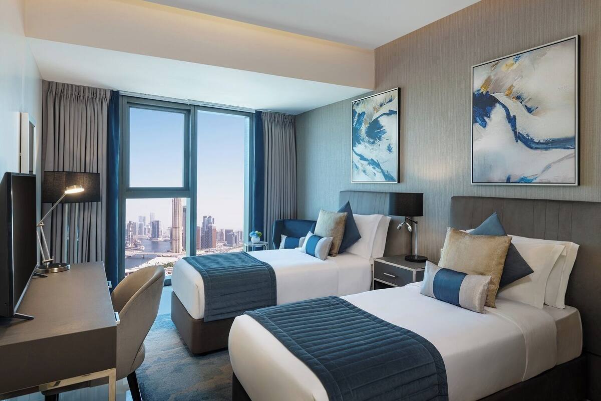 A hotel room with two beds, a desk, and a large window offering a city view.