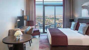 A modern hotel room with a large bed, a desk, and a view of a cityscape through floor-to-ceiling windows.