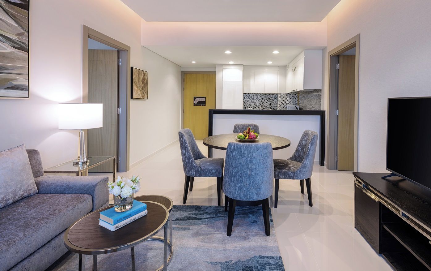 A modern living room with a sofa, coffee table, and TV. There is a dining area with a table and chairs. The room has a kitchen in the background.