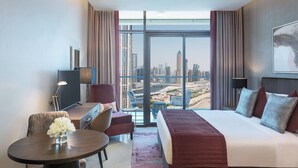 A hotel room with a large bed, a desk, a chair, and a view of the cityscape through floor-to-ceiling windows. - Damac Maison Aykon City Dubai (Dubai)