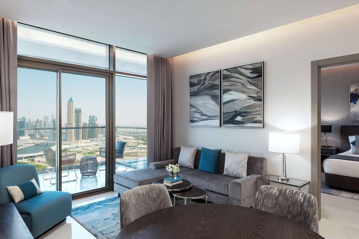 A modern hotel room with a large window offering a city view, a sofa, a dining table, and a bed.