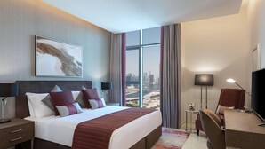 A modern hotel room with a large bed, a desk, a chair, a TV, and a city view through large windows. - Damac Maison Aykon City Dubai (Dubai)
