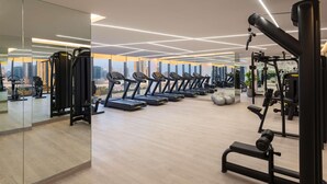 Un gimnasio moderno con cintas de correr, máquinas de pesas y un espejo grande que refleja el paisaje urbano.