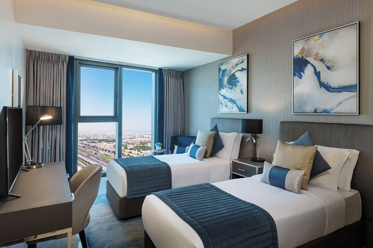 A hotel room with two beds, a desk, a chair, and a large window offering a city view.